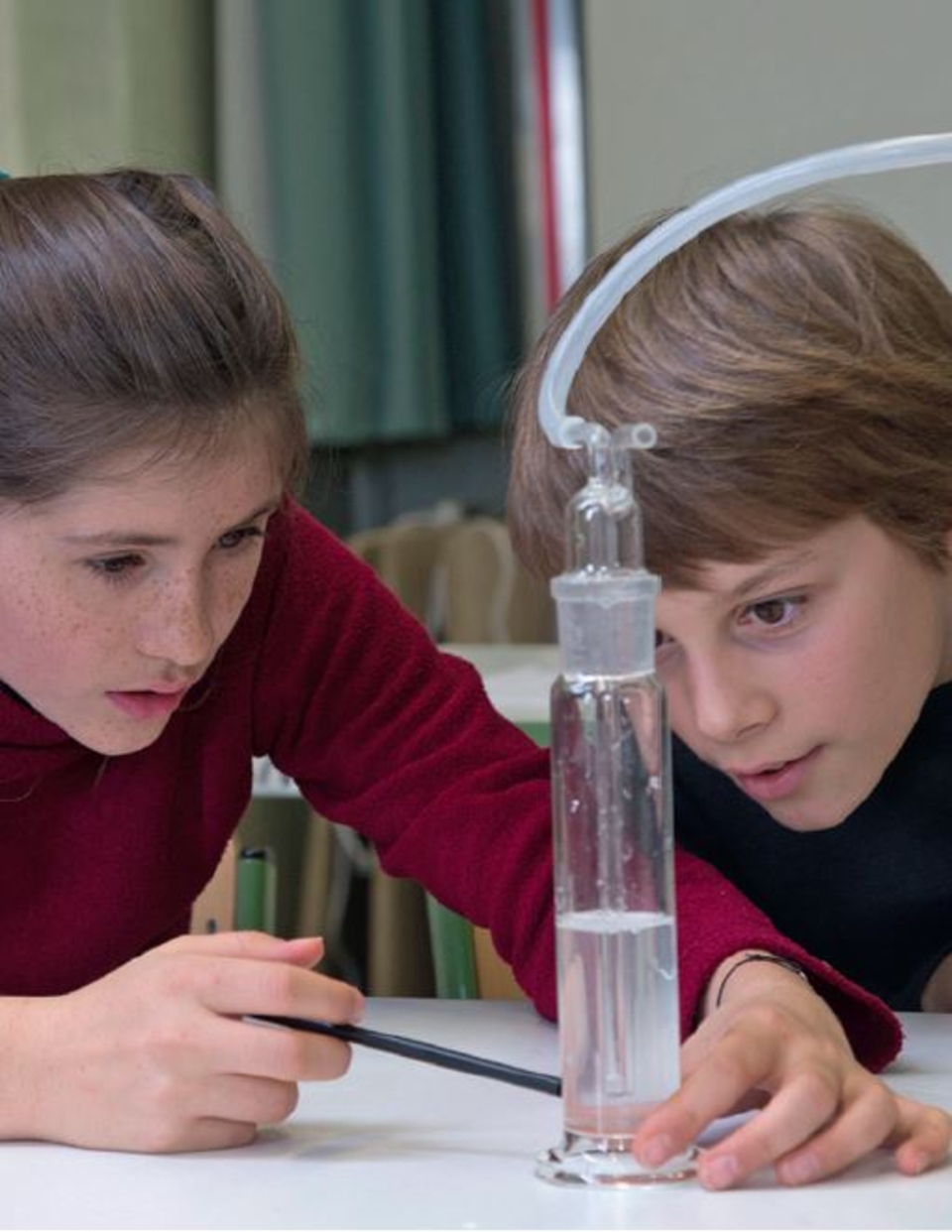 Das Bild zeigt zwei Kinder, die konzentriert an einem Experiment mit einemZylindermessger&auml;t arbeiten. Sie beobachten den Wasserstand und verwenden ein spezielles Ger&auml;t, um den Vorgang zu steuern, was auf ein naturwissenschaftliches Experiment hinweist.