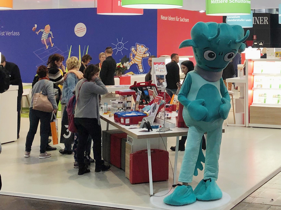 Das Bild zeigt einen Messestand f&uuml;r Experimentierbedarf mit mehreren Erwachsenen, die an einem Tisch interessiert Produkte betrachten. Im Vordergrund steht eine Person in einem gr&uuml;nen Kost&uuml;m, die das Interesse der Besucher auf sich zieht.