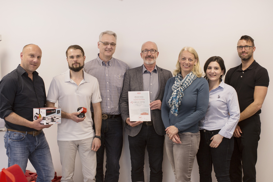 Das Bild zeigt eine Gruppe von sechs Personen, die zusammen stehen und verschiedene Produkte sowie ein Zertifikat in der Hand halten. Sie scheinen an einem Projekt im Bereich Experimentierbedarf f&uuml;r das Bildungswesen beteiligt zu sein.
