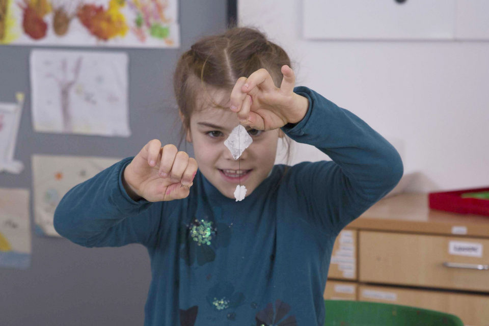 Ein M&auml;dchen h&auml;lt stolz ein kleines Experiment in der Hand, das aus einem transparenten, gefalteten Material und etwas Wei&szlig;em besteht. Im Hintergrund sind Zeichnungen von anderen Kindern zu sehen, die eine kreative Umgebung schaffen.