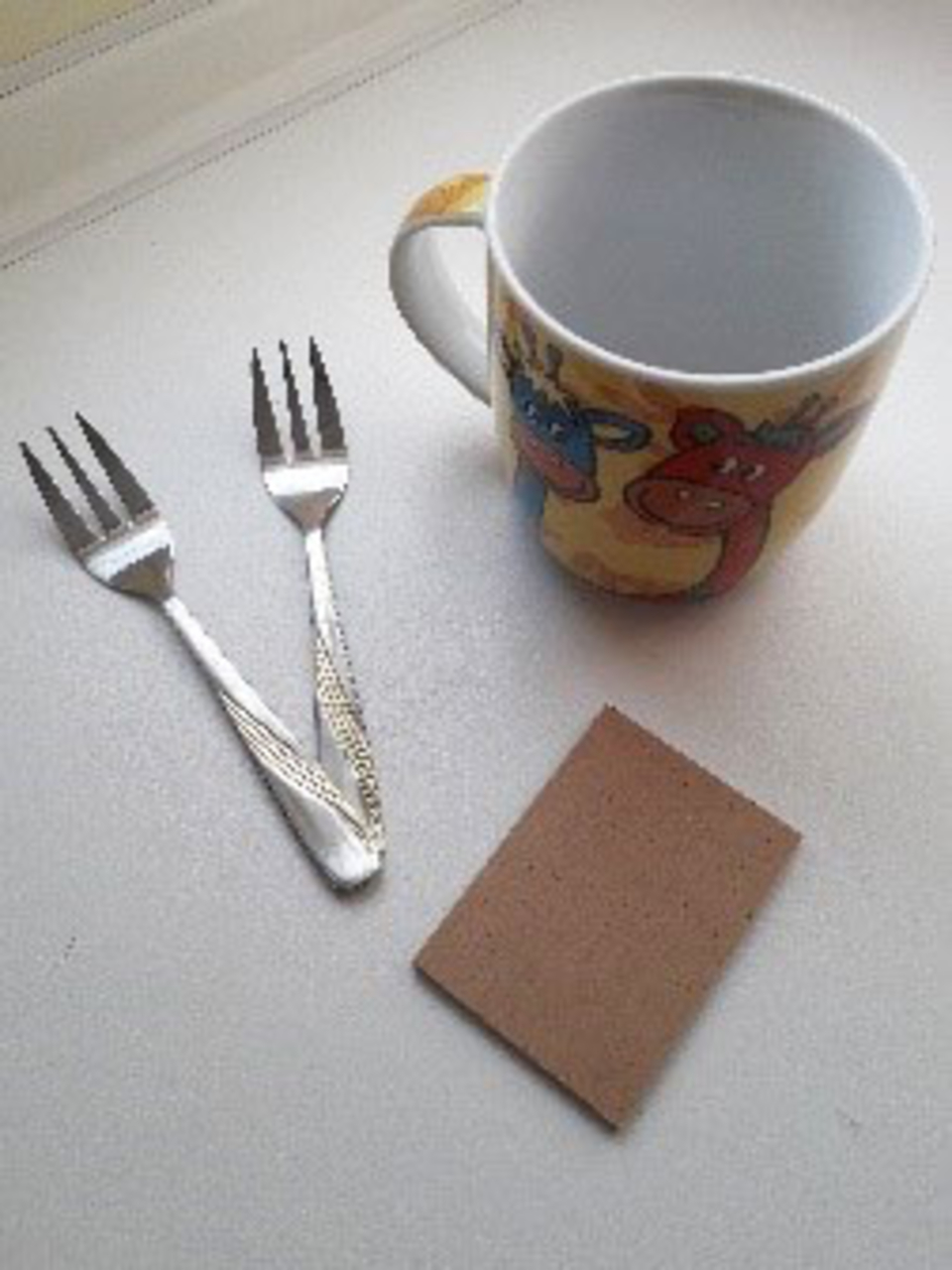 Das Bild zeigt eine bunte Tasse mit Tiermotiven, zwei kleine Gabeln und ein rechteckiges St&uuml;ck Pappe. Diese Objekte k&ouml;nnten f&uuml;r verschiedene Bildungsaktivit&auml;ten oder Experimentierprojekte im schulischen Kontext verwendet werden.