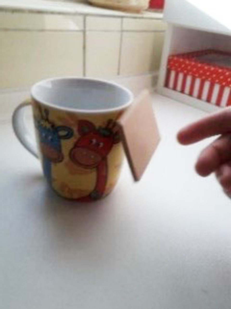 Das Bild zeigt eine bunte Tasse mit Tiermotiven, die auf einem Tisch steht. An der Tasse rechts befindet sich ein kleines Holzst&uuml;ck, das zu schweben scheint, daneben ist noch eine Hand zu erkennen, die das Holzst&uuml;ck nicht festh&auml;lt.