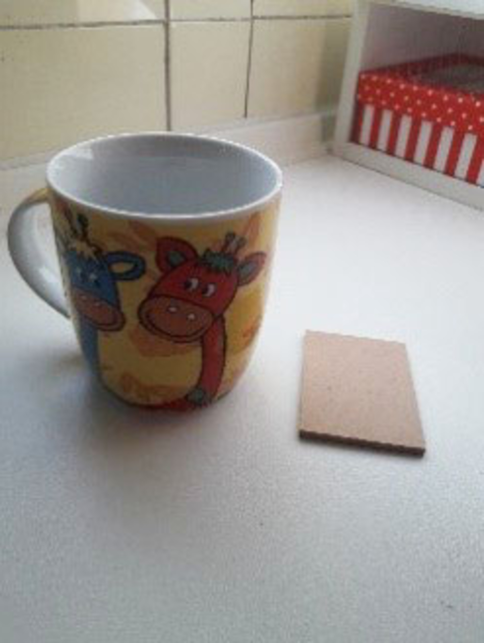 Auf dem Bild ist eine bunte Tasse mit Kuh-Motiven und ein rechteckiges St&uuml;ck Pappe zu sehen. Der Hintergrund zeigt eine helle Oberfl&auml;che, die in einem p&auml;dagogischen Kontext genutzt werden k&ouml;nnte.