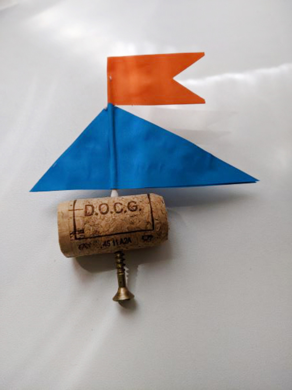 Das Bild zeigt ein einfaches Bootmodell, das aus einem Korken als Rumpf und bunten Papierdreiecken als Segeln besteht. Die blaue und orangefarbene Flagge verleihen dem Modell einen ansprechenden Look f&uuml;r experimentelle Zwecke im Bildungsbereich.