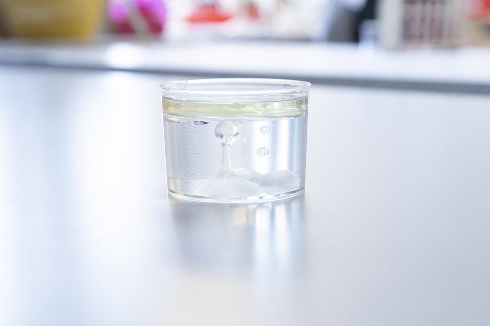 Das Bild zeigt ein Becherglas mit einer klaren Fl&uuml;ssigkeit und einer schaumigen Phase an der Unterseite. Es sind kleine Luftblasen zu sehen, die im Glas aufsteigen, w&auml;hrend sich eine gelbliche &Ouml;lschicht an der Oberfl&auml;che bildet.