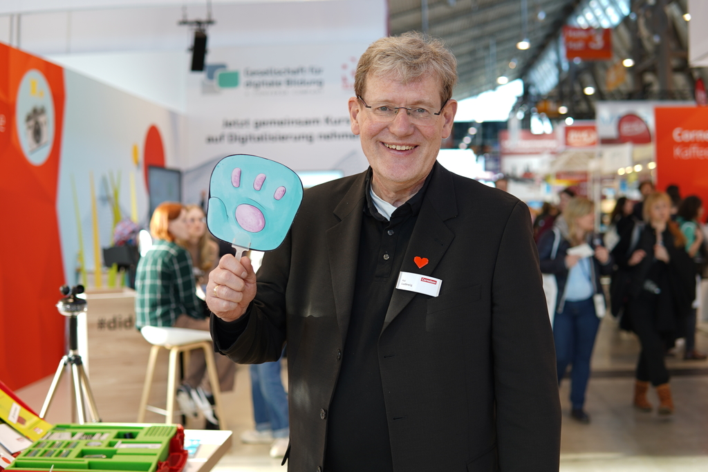 Das Bild zeigt einen l&auml;chelnden Mann in einem schwarzen Blazer, der ein plakatiertes Hand-Symbol h&auml;lt. Im Hintergrund sind Menschen und St&auml;nde einer Bildungsmesse zu sehen, was auf eine Veranstaltung f&uuml;r experimentelle Lernmaterialien hindeutet.