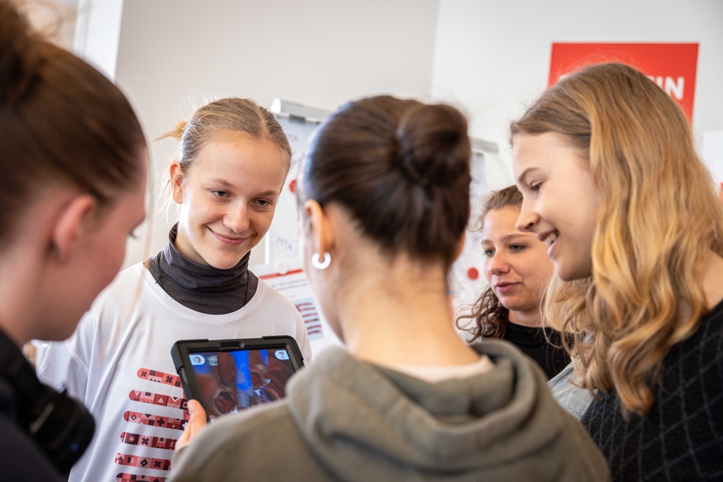 Auf dem Bild sind mehrere M&auml;dchen zu sehen, die fasziniert auf ein Tablet schauen, das mithilfe von Augmented Reality das K&ouml;rperinnere zeigt. Ein M&auml;dchen hat das Magic T-Shirt an und l&auml;chelt, w&auml;hrend die anderen aufmerksam dem Inhalt auf dem Bildschirm fo
