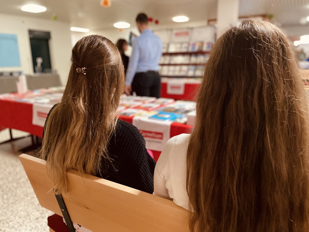 Das Bild zeigt zwei Sch&uuml;lerinnen, die auf einer Bank sitzen und sich den Cornelsen-Stand mit B&uuml;cher- und Experimentiermaterialien im Hintergrund anschauen. Dieser Stand ist im Rahmen einer Veranstaltung f&uuml;r das deutsche Bildungswesen aufgebaut.
