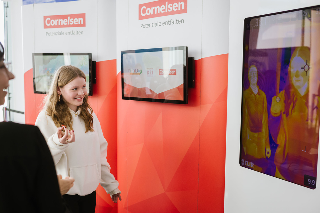 Girls_Day_2024_Schuelerin_W&auml;rmebildkamera / Ein M&auml;dchen in einem wei&szlig;en Hoodie l&auml;chelt und zeigt auf ein Display mit einer W&auml;rmebildaufnahme. Das Display zeigt das Cornelsen-Logo und den Slogan "Potenziale entfalten". Im Hintergrund befinden sich weitere 