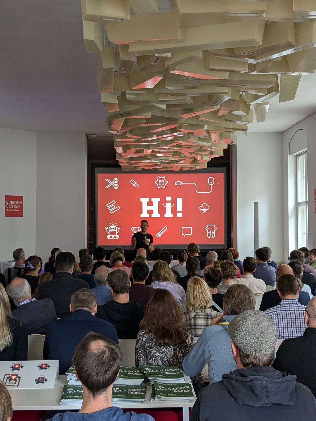 Das Bild zeigt eine Veranstaltung in einem modernen Raum, wo ein Sprecher vor einem gro&szlig;en Bildschirm mit der Begr&uuml;&szlig;ung "Hi!" pr&auml;sentiert. Im Vordergrund sind Teilnehmer zu sehen, w&auml;hrend auf Tischen verschiedene Materialien und Informationsbl&auml;tter liegen