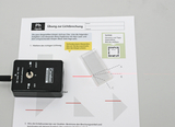 Das Bild zeigt ein Experimentiergerät zur Lichtbrechung, das aus einem Lasermodul und einer Glasplatte besteht, zusammen mit einer Instruktionsseite. Die Seite enthält Aufgaben zur Untersuchung des Lichtwegs und zur Bestimmung von Brechungswinkeln.