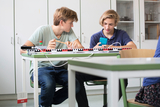 Das Bild zeigt zwei Schüler in einem Klassenzimmer, die an einem Experimentiergerät arbeiten. Der Junge links konzentriert sich auf das Gerät, während der andere mit einem Smartphone beschäftigt ist.