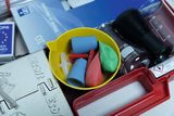 Das Bild zeigt eine gelbe Schüssel mit verschiedenen Büro- und Experimentiermaterialien, darunter bunte Gummibälle, ein Spritzgerät und mehrere Messbecher. Im Hintergrund sind weitere Utensilien zu sehen, die für experimentelle Anwendungen im Bildungsbere
