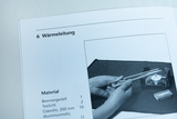 Das Bild zeigt eine Anleitung zum Experimentieren mit Wärmeleitung, in der zwei Glasrohre in den Händen gehalten werden. Im Hintergrund sind ein Teelicht und ein Brennergestell auf einer Arbeitsfläche zu sehen.