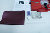 Das Bild zeigt eine Sammlung von Experimentiermaterialien, darunter ein lila Stück Stoff, einige Reagenzgläser, eine Kerze, ein Streichholzbuch und eine Metallbox. Diese Materialien sind in einem roten Kasten angeordnet, begleitet von einem Handbuch mit A