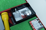 Das Bild zeigt einen Experimentierkasten mit verschiedenen Werkzeugen und Materialien, darunter eine Taschenlampe, Batterien und weitere kleine Utensilien. Die Aufbewahrungseinheit ist in einem roten Kasten mit grünem Schaumstoff-Innenleben gestaltet.