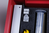 Das Bild zeigt eine rote Aufbewahrungsbox mit verschiedenen Experimentiermaterialien. Darin befinden sich ein graues Halterungselement, ein Messzylinder sowie weitere Teile, die für wissenschaftliche Experimente im Bildungsbereich eingesetzt werden können