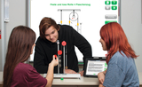 Das Bild zeigt drei SchülerInnen, die an einem Experiment mit Rollen und einem Flaschenzug arbeiten. Im Hintergrund ist eine digitale Tafel zu sehen, die die Konzepte von festen und losen Rollen erklärt.