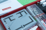 Das Bild zeigt einen Experimentierkasten aus einem Online-Shop für Bildung, der verschiedene Materialien für naturwissenschaftliche Experimente enthält. Sichtbar sind eine Anleitung, ein metallischer Verbindungsträger und Werkzeugutensilien, die zum Exper