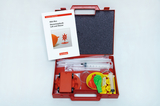 Das Bild zeigt eine rote Box mit Experimentiermaterialien zur Elementarphysik, die sich auf Luft und Wasser konzentrieren. In der Box befinden sich verschiedene Utensilien wie Spritzen, Luftballons und ein Experimentierheft, die für Schulprojekte verwende
