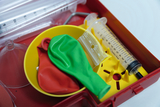 Das Bild zeigt einen Experimentierkasten mit verschiedenen Materialien für den Unterricht, darunter rote und grüne Ballons, eine Spritze sowie mehrere transparente Röhren. Die Gegenstände sind in einem roten Kasten angeordnet, der für praktische Versuche 