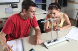 Das Bild zeigt zwei Schüler, die an einem Experimentiergerät arbeiten, um physikalische Konzepte zu erkunden. Im Fokus steht eine Apparatur mit Gewichten und einem Messzylinder, während beide aufmerksam Notizen machen und diskutieren.