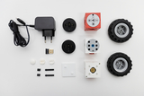 Das Bild zeigt eine Auswahl an Einzelteilen für ein Experimentierset, darunter verschiedene Würfel, Räder, einen Netzadapter und USB-Zubehör. Die Teile scheinen für den Zusammenbau eines technischen Modells gedacht zu sein, das Programmierung oder Robotik