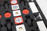 Das Bild zeigt eine Anordnung von verschiedenen modularen Bauteilen in einer Schaumstoff-Einlage, die für experimentelle Lernzwecke verwendet werden. Zu sehen sind rote und weiße Elemente, darunter Räder und Steuerungsplatinen, die interaktive Projekte er