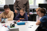 Das Bild zeigt eine Gruppe von Schülern, die an einem Tisch sitzen und sich mit einem kleinen Roboter beschäftigen. Sie wirken interessiert und lachen, während ein Erwachsener im Hintergrund steht und ihnen möglicherweise Anweisungen gibt.