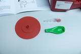 Das Bild zeigt ein Experimentierset für Bildungseinrichtungen, bestehend aus einer roten Basisplatte, einem grünen Ballon und einem Verbindungsteil. Neben den Materialien liegt ein Handbuch, das Anweisungen zur Durchführung eines Luftexperiments enthält.