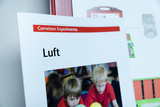 Das Bild zeigt ein Arbeitsblatt mit dem Titel „Luft“ aus dem Cornelsen Experimenta Material, das in Bildungsbereichen verwendet wird. Darunter ist ein Foto von zwei Kindern, die mit bunten Bällen spielen und experimentieren.