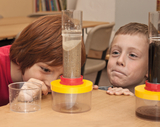Auf dem Bild sind zwei Kinder zu sehen, die fasziniert auf ein Experimentierset schauen, das anscheinend verschiedene Schichten von Materialien zeigt. Das Set besteht aus transparenten Behältern, die unterschiedlich gefärbte Substanzen enthalten, und weck