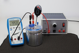 Das Bild zeigt ein Experimentierset mit einem Multimeter, das zur Messung der Stromstärke dient, und einer Stromquelle, die sowohl Gleich- als auch Wechselstrom bereitstellen kann. In der Mitte befindet sich ein Behälter, der vermutlich für Experimente ge