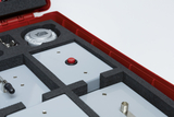 Das Bild zeigt ein rotes Zubehörset mit verschiedenen Experimentiermodulen, die in schaumstoffgepolsterten Fächern angeordnet sind. Zu sehen sind unter anderem ein großer roter Knopf und metallische Elemente, die für Experimentierzwecke genutzt werden kön