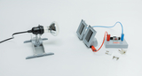 Das Bild zeigt eine Experimentierausrüstung, die aus einer Lampenhalterung mit Glühlampe und einer Versuchsplatine mit elektrischen Komponenten besteht. Daneben liegen verschiedene Stecker, die für elektrische Verbindungen genutzt werden können.