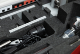 Das Bild zeigt einen Koffer mit verschiedenen Experimentierutensilien, die in schaumstoffgepolsterten Fächern organisiert sind. Zu sehen sind unter anderem ein Kabel, eine Lampe und metallische Teile, die für Bildungszwecke genutzt werden können.