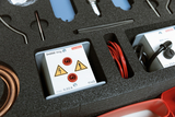 Das Bild zeigt eine Box mit wissenschaftlichen Experimentiergeräten, darunter zwei Geräte mit elektrischen Anschlüssen und Warnsymbolen. Zudem sind verschiedene Kabel, sowie weitere Instrumente in Schaumstoff-Formteilen untergebracht, die zur sicheren Aufbewahrung dienen.