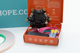 Das Bild zeigt ein Experimentier-Set, das einen Calliope mini Mikrocontroller sowie Zubehör wie Kabel und Batterien enthält. Die orangefarbene Verpackung bietet Anleitungen zur Programmierung und Nutzung des Calliope mini für Bildungszwecke.