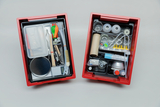 Das Bild zeigt zwei rote Boxen, die mit verschiedenen Experimentiermaterialien gefüllt sind. Die linke Box enthält Werkzeuge, Behälter und Materialien, während die rechte Box hauptsächlich Gläser, Rohre und weitere Chemikalien benötigt für Experimente im Bildungsbereich umfasst.