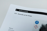 Das Bild zeigt eine Seite aus einem Unterrichtsleitfaden, die mit dem Thema "Verbrennung" und speziell mit "Eisenwolle auf der Waage" befasst ist. Außerdem sind Sicherheitszeichen und Hinweise zu Materialien und Schutzmaßnahmen abgebildet.