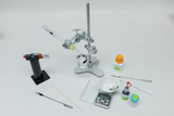 Das Bild zeigt eine Experimentierausstattung, die verschiedene Laborgeräte und Chemikalien umfasst. Zu sehen sind unter anderem ein Bunsenbrenner, eine präzise Waage und Glaswaren, die für chemische Versuche genutzt werden können.