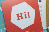 Das Bild zeigt eine quadratische blaue und rote Karte, auf der in großen, roten Buchstaben "Hi!" steht. Im Hintergrund sind weitere farbige Materialien und Texte sichtbar, die auf Experimentierbedarf für das Bildungswesen hinweisen.