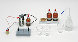 Das Bild zeigt eine Laborausrüstung, die verschiedene Chemikalien in braunen Glasflaschen beinhaltet, darunter Essigsäure, Salzwasser und Schwefelsäure, sowie zwei Messbecher. Zusätzlich sind ein Batterie-Stromkreis und eine Pipette zur Handhabung der Flüssigkeiten abgebildet.