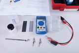 Das Bild zeigt verschiedene Experimentiermaterialien, darunter ein Multimeter, ein Becher, metallische Platten, eine Lötzange und ein Löffel. Diese Utensilien sind typisch für chemische oder physikalische Experimente im Bildungsbereich.