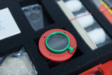 Das Bild zeigt einen Kasten mit verschiedenen Experimentiermaterialien, darunter ein rot-grünes Linsengerät und mehrere Komponenten. Die Anordnung auf schwarzem Schaumstoff hebt die Einzelteile hervor, die für Bildungs- oder Experimentierzwecke genutzt werden können.