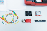 Das Bild zeigt verschiedene Experimentiermaterialien, darunter bunte Kabel, eine Glocke, eine Batteriehalterung, ein Schalter und Batterien. Diese Materialien sind Teil eines Sets für elektrische Experimente im Bildungsbereich.