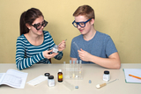Das Bild zeigt zwei Schüler, die gemeinsam an einem Experiment arbeiten. Sie haben verschiedene Chemikalien und Laborausrüstung auf dem Tisch, während sie konzentriert und fröhlich interagieren.