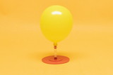 Das Bild zeigt einen gelben Ballon, der auf einer roten Basis steht und durch einen transparenten Schlauch verbunden ist. Die gesamte Komposition hat einen fröhlichen, einladenden Look vor einem einfarbigen gelben Hintergrund.
