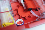 Das Bild zeigt ein rotes Kunststoff-Kit mit verschiedenen Experimentiermaterialien, einschließlich Röhren und eines speziellen Halters. Es ist für den Einsatz im Bildungsbereich konzipiert, um wissenschaftliche Experimente im Unterricht durchzuführen.