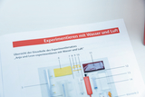 Das Bild zeigt eine Übersicht der Einzelteile eines Experimentiersatzes mit dem Titel „Anja und Leon experimentieren mit Wasser und Luft“. Die Abbildung listet verschiedene Komponenten auf, die für die Durchführung von Experimenten benötigt werden.